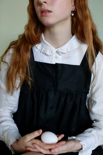 Female model wearing a long sleeve white linen shirt with a Peter Pan collar under a black sleeveless jumper 