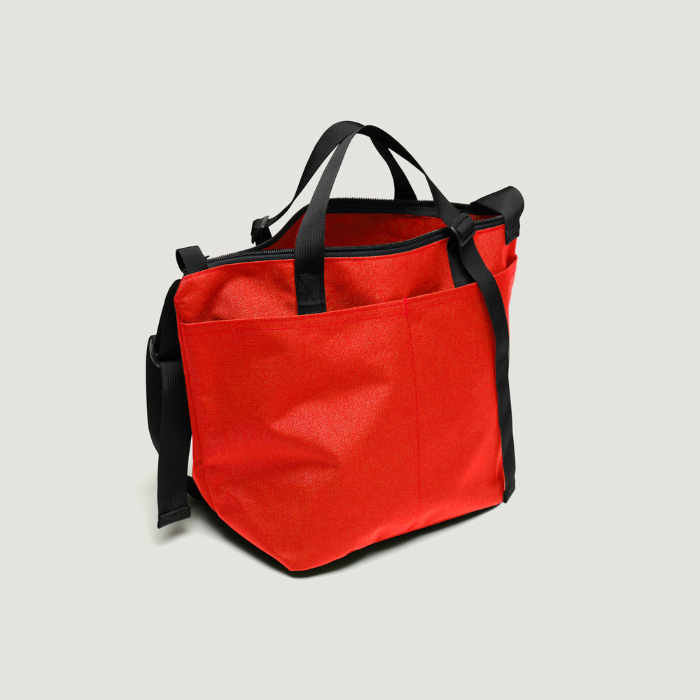 Reddish-orange tote bag with exterior pockets, black handle & shoulder strap, and  zipper closure (back view)