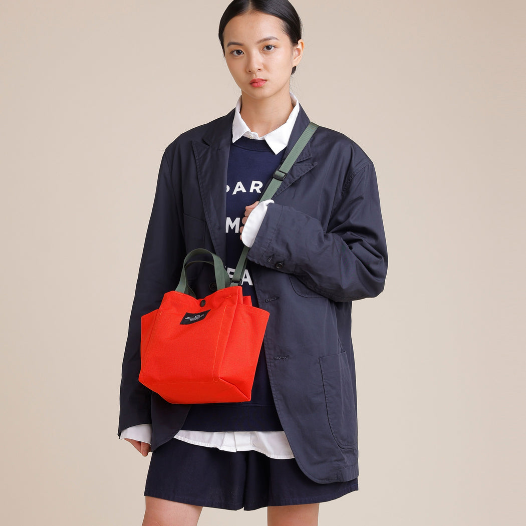 Female model wearing a reddish-orange small tote with cross body olive green adjustable shoulder strap