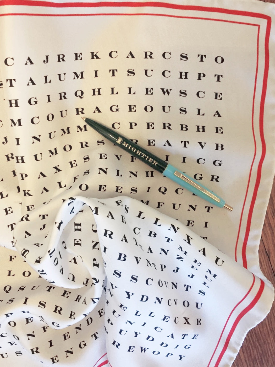 Close-up of white silk scarf with red border and black word search letters with pen sitting atop