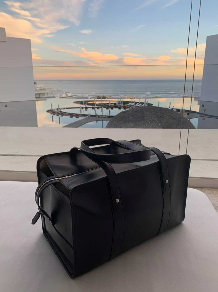 Three-quarter view of black leather travel bag with leather straps & zipper closure