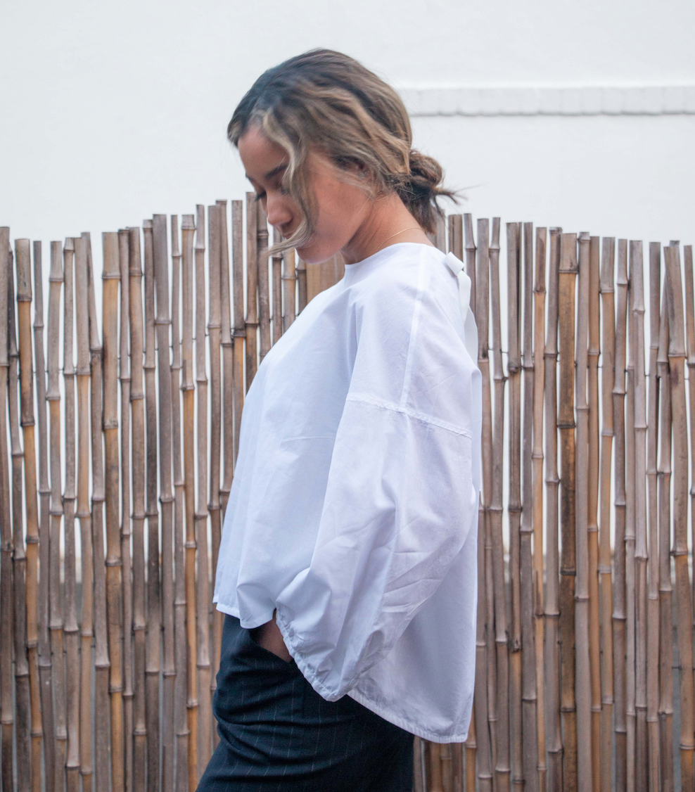 Side view: Female model wearing a white semi-sheer popover shirt with puffy long sleeves and rounded neck and hemline