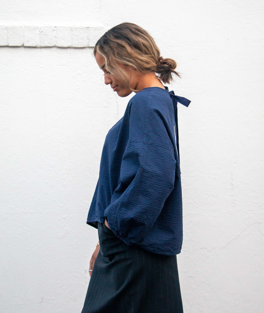 Side view: Model wearing dark blue popover shirt with long sleeves and grosgrain ribbon tie back closure and striped crinkle