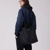 Male model wearing a large black tote across his body with black nylon adjustable shoulder straps