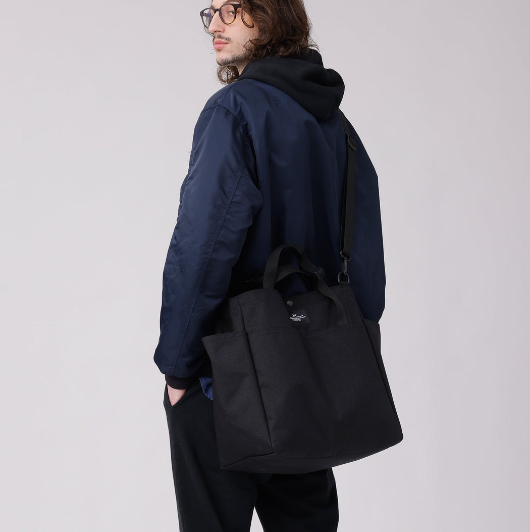 Male model wearing a large black tote across his body with black nylon adjustable shoulder straps