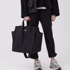 Model holding a large black tote bag with exterior pockets and adjustable handles and shoulder strap