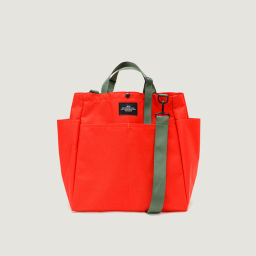 Large reddish-orange tote bag with snap closure, exterior pockets, and olive green handles & shoulder strap (front view)