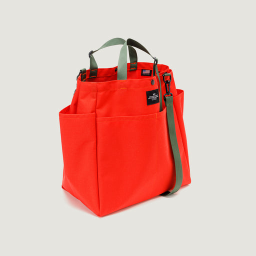 Large reddish-orange tote bag with snap closure, exterior pockets, and olive green handles & shoulder strap (3/4 view)