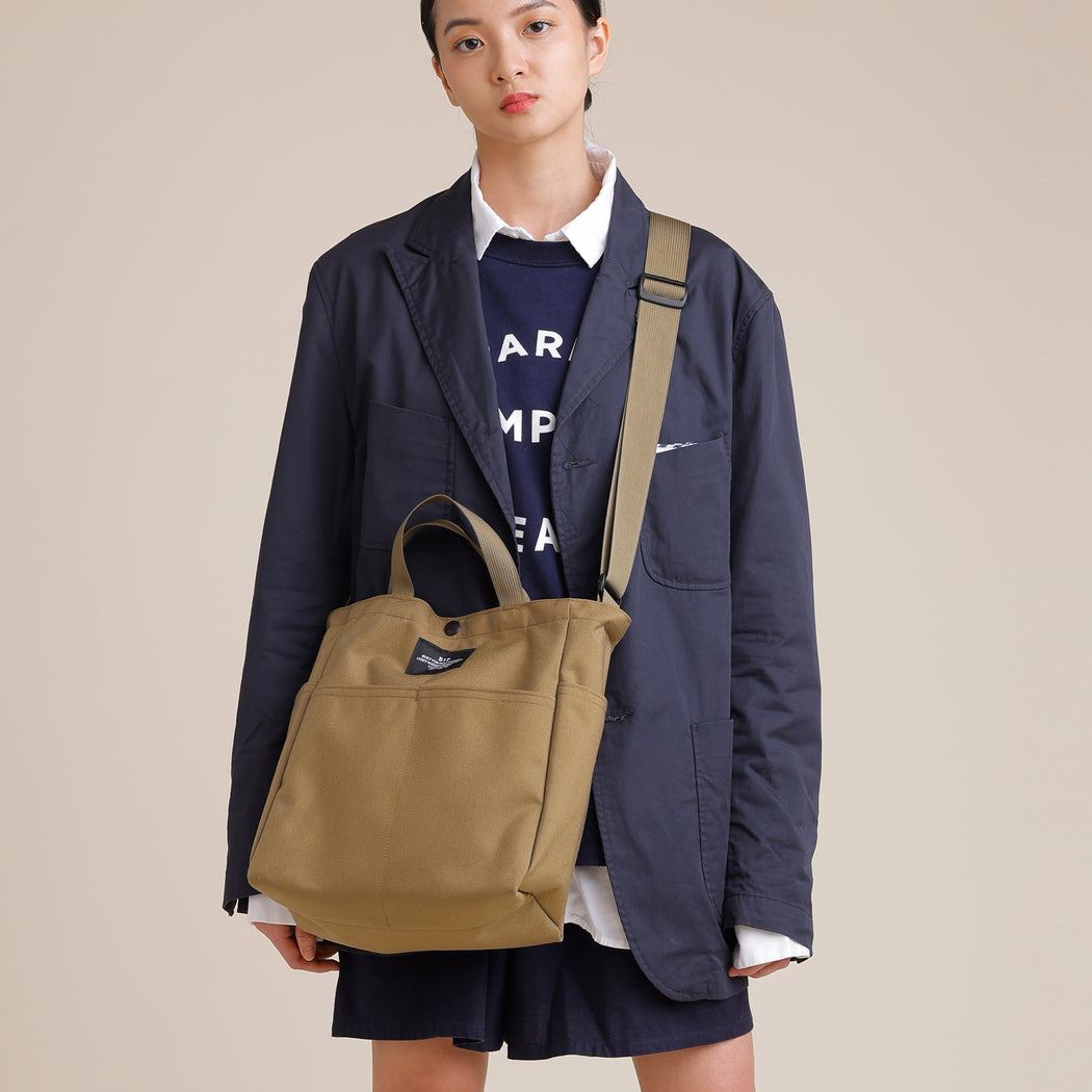 Female model wearing khaki colored canvas tote with 4 exterior pockets, snap closure, handles & shoulder strap
