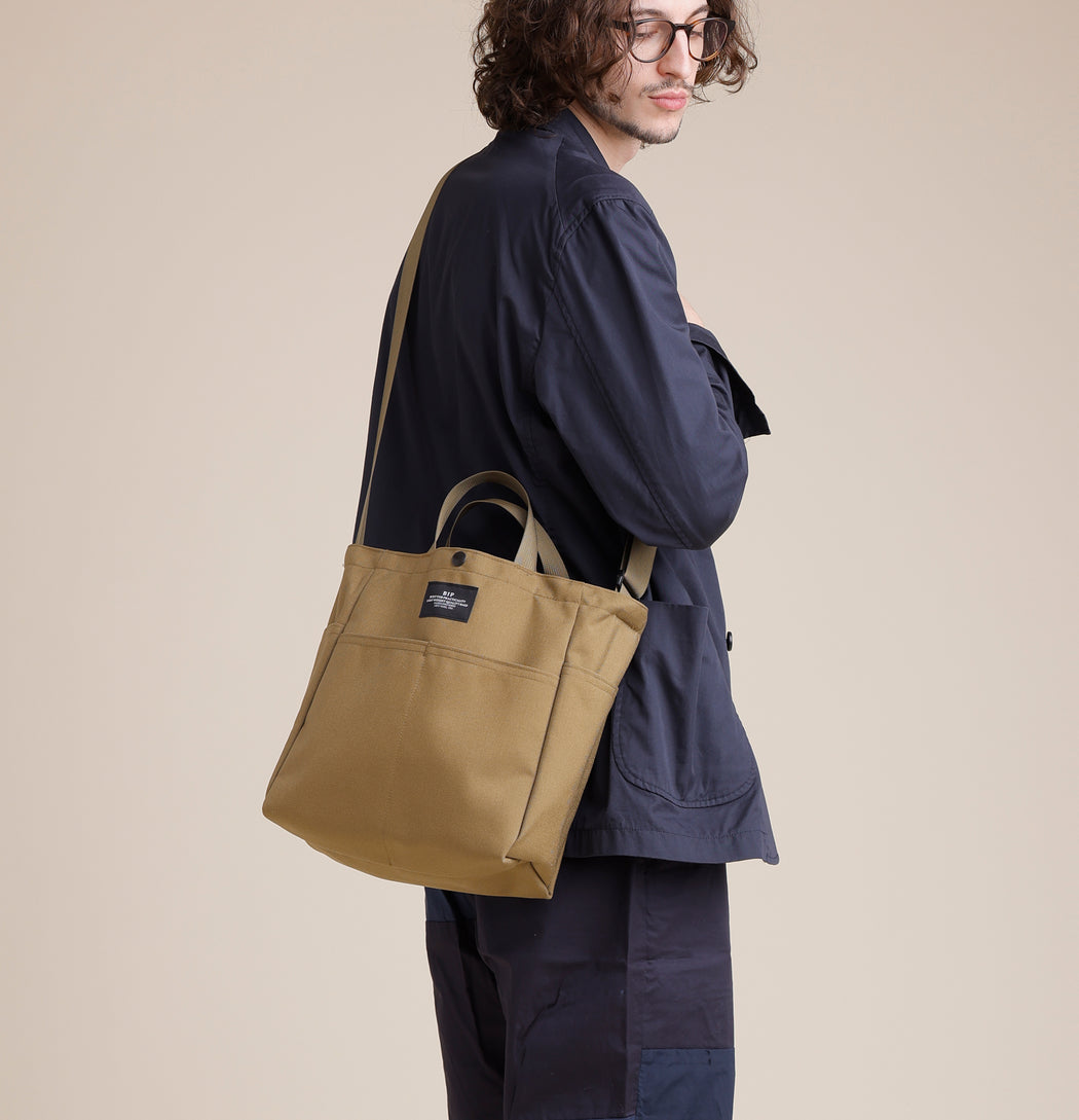 Male model wearing khaki colored canvas tote with 4 exterior pockets, snap closure, handles & shoulder strap