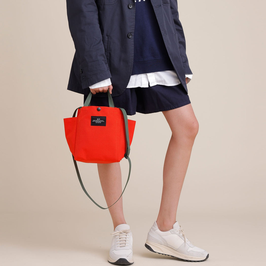 Female model holding a reddish-orange small tote by the handles down by her side