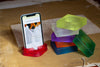 Red silicone square bracelet being used as a phone holder next to a stack of rainbow bracelets