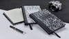 Three black & white composition notebooks (1 open, 2 closed) next to a pen and a camera