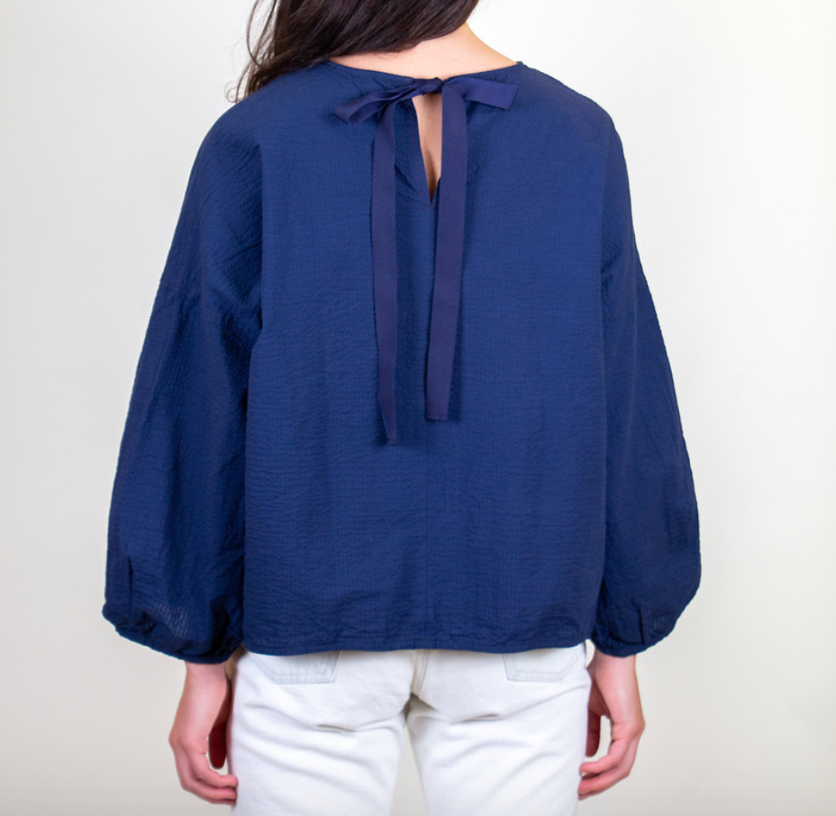 Back view: Model wearing dark blue popover shirt with long sleeves and grosgrain ribbon tie back closure and striped crinkle