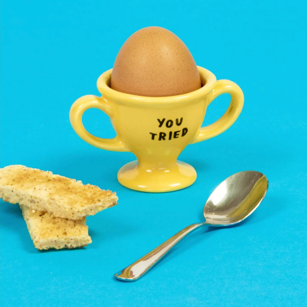 Brown egg sitting inside a yellow double-handled ceramic egg cup reading "You Tried" in black (alongside 2 wafers and a spoon)