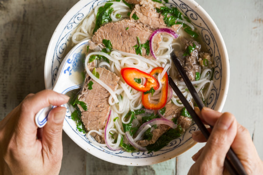 A bowl of pho with 2 hands holding a spoon and a pair of chopsticks