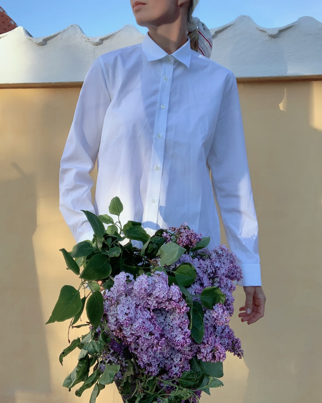 Model wearing white button down collar shirt, holding a bouquet of lilacs