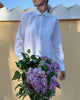 Model wearing white button down collar shirt, holding a bouquet of lilacs